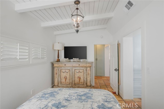Bedroom with floral bedding, plantation shutters, exposed ceiling beams, and an antique cabinet with TV.