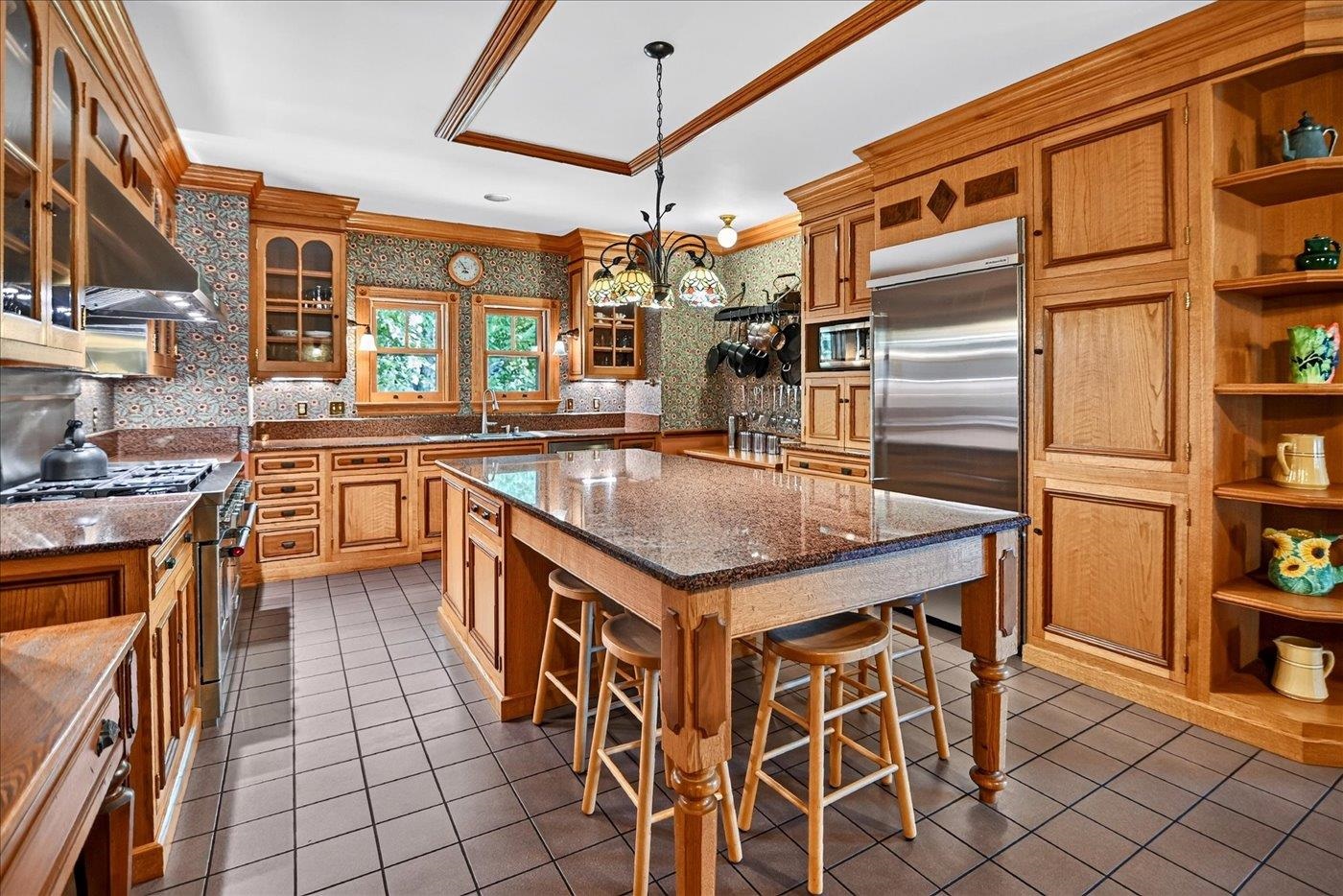 Kitchen with honey-toned wood cabinetry, patterned wallpaper, and a large granite island with bar stools.
