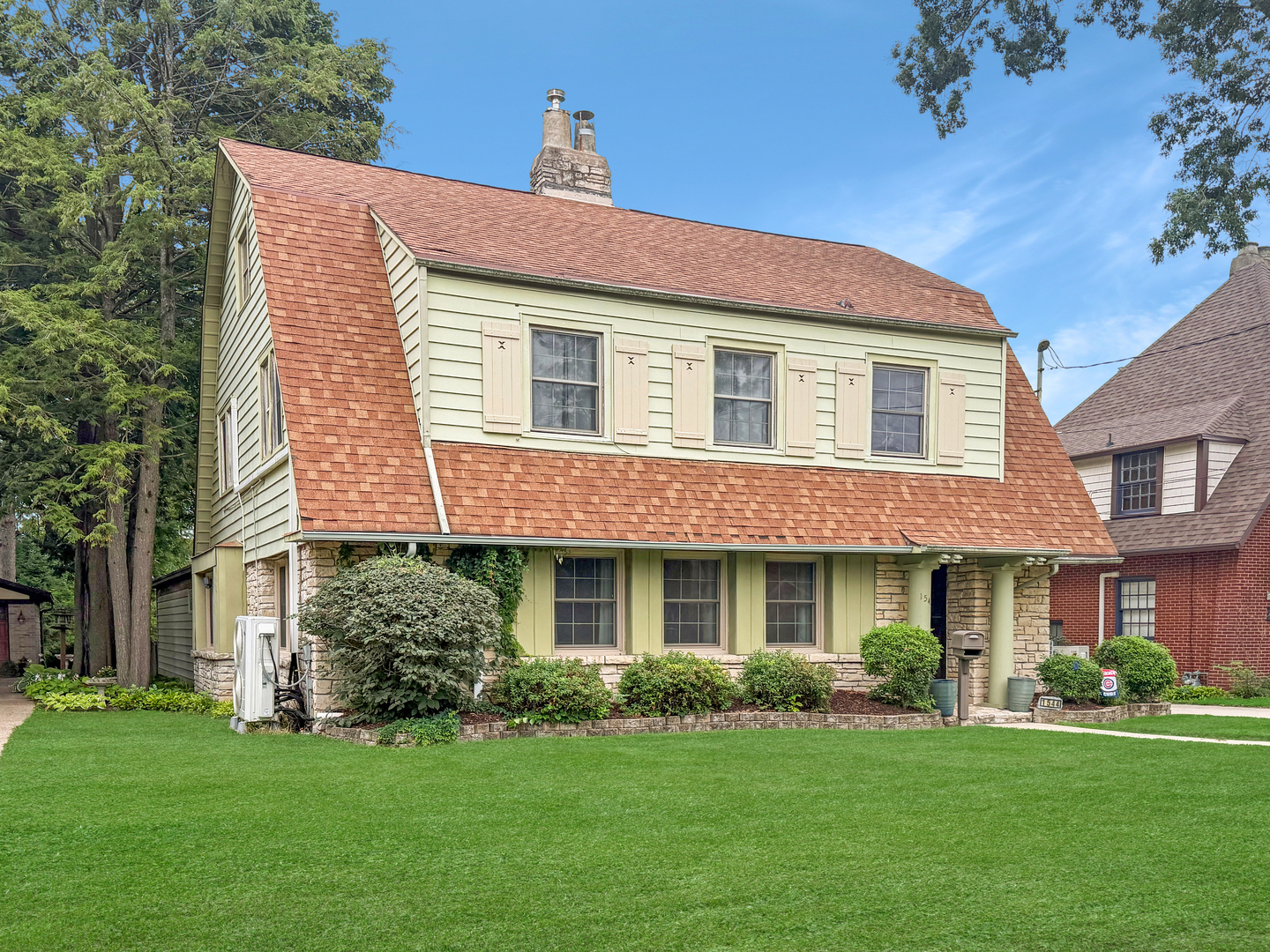 Pale green two-story house with a broad reddish-brown roof, stone lower walls, and a manicured lawn.
