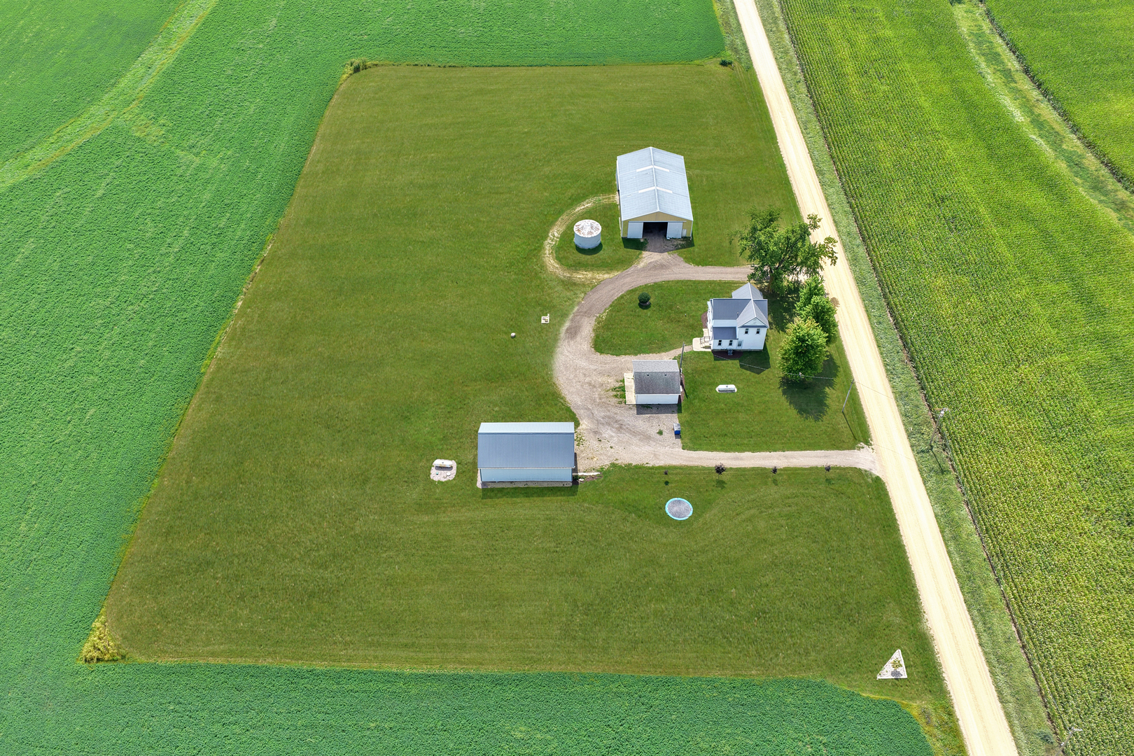 Aerial view of a rural home and outbuildings amid crop fields, with a gravel drive and round pool.