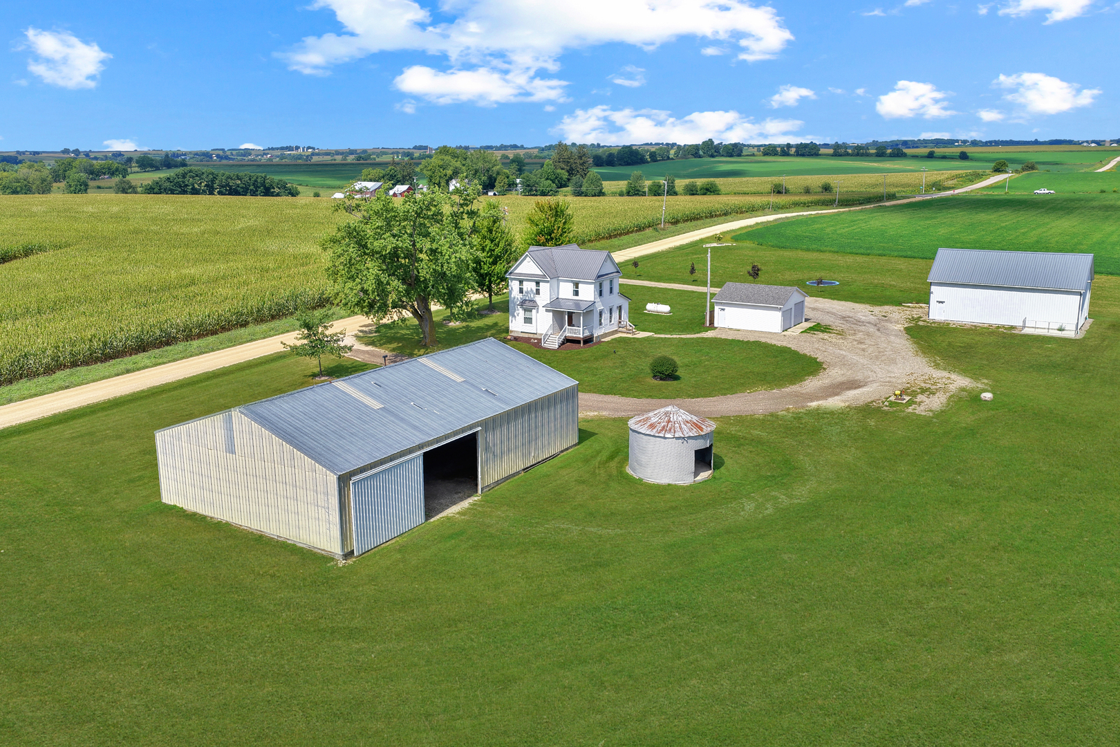 Aerial view of a white farmhouse, multiple metal outbuildings, and a silo surrounded by lawn and cornfields.