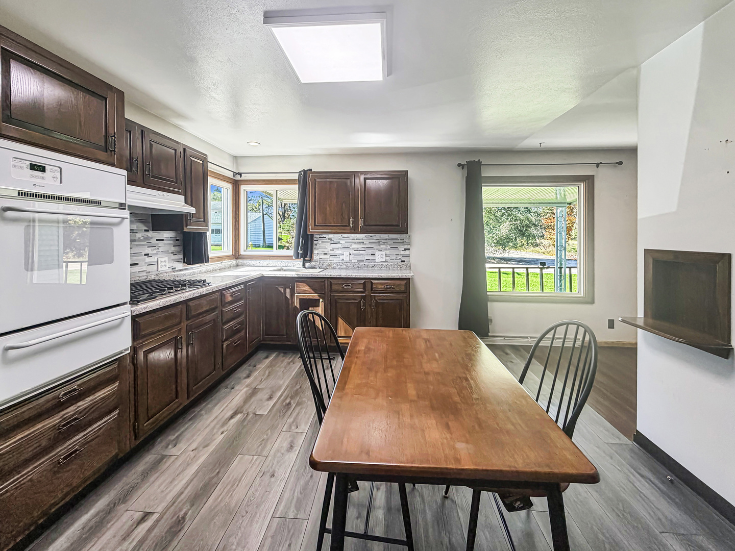 Kitchen and dining area with dark cabinets, white appliances, and a wooden table by large windows.