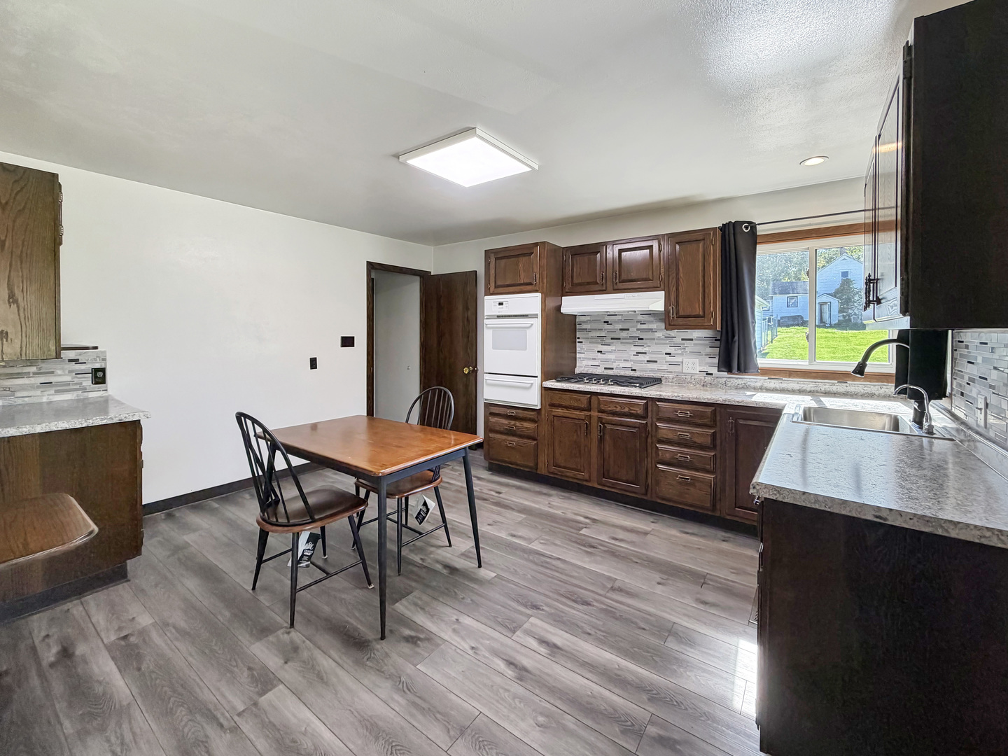 Kitchen with dark wood cabinets, gray countertops, and a dining table beside a sunny window.