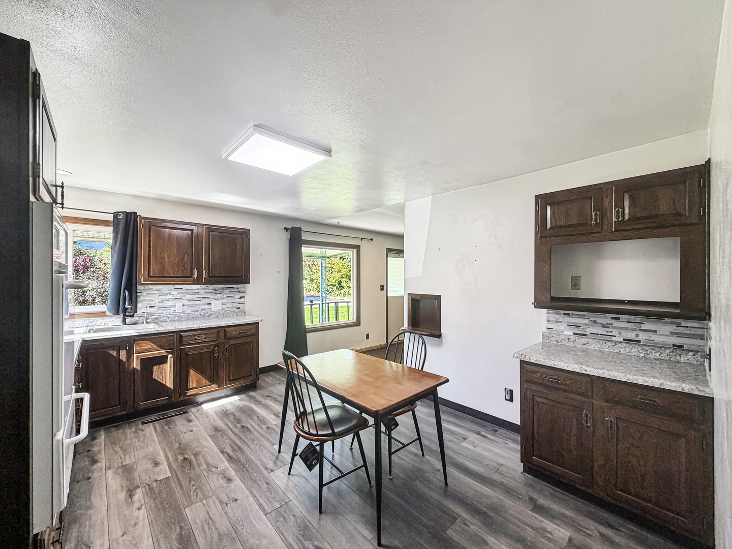 Kitchen and dining area with dark wood cabinets, gray counters, and a table beside a leafy window.