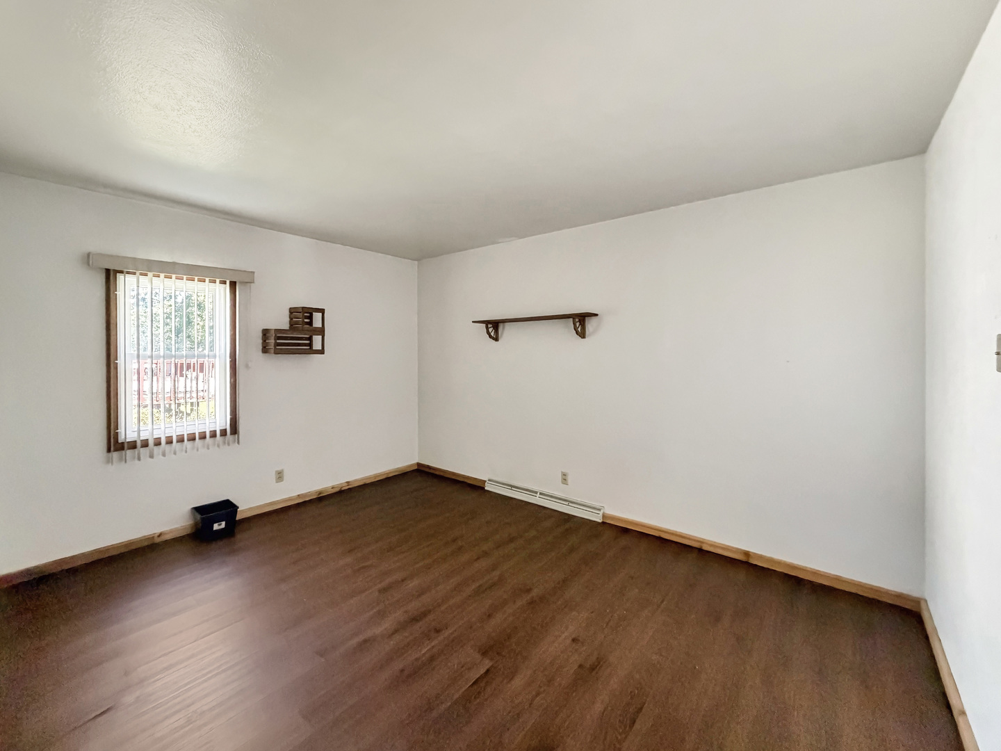 Vacant room with dark wood floors, a window with vertical blinds, and small wall shelves.