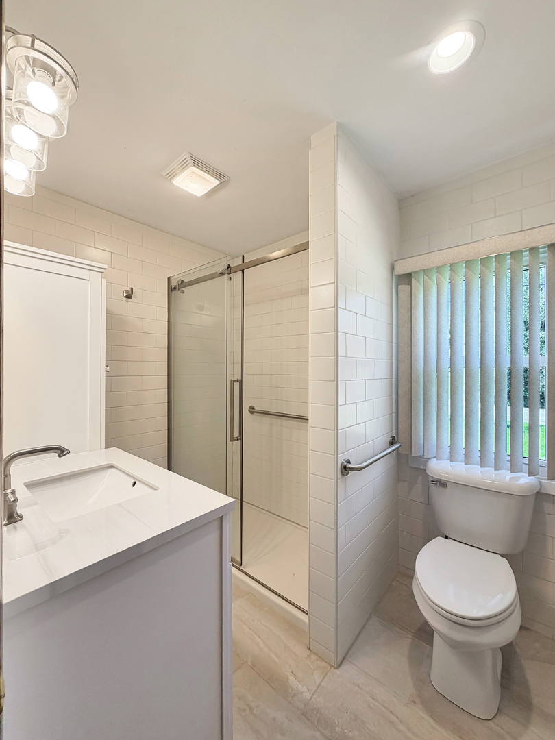 Bathroom with a glass-enclosed shower, white tiled walls, and a toilet beneath vertical blinds.
