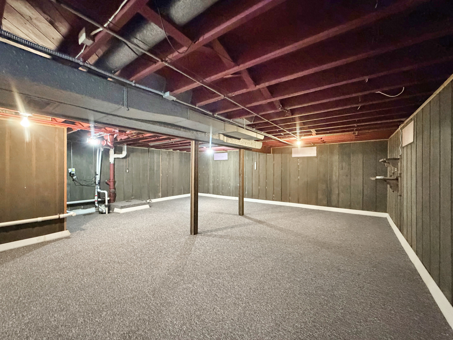 Basement with gray carpet, exposed ceiling joists, support posts, and visible plumbing.