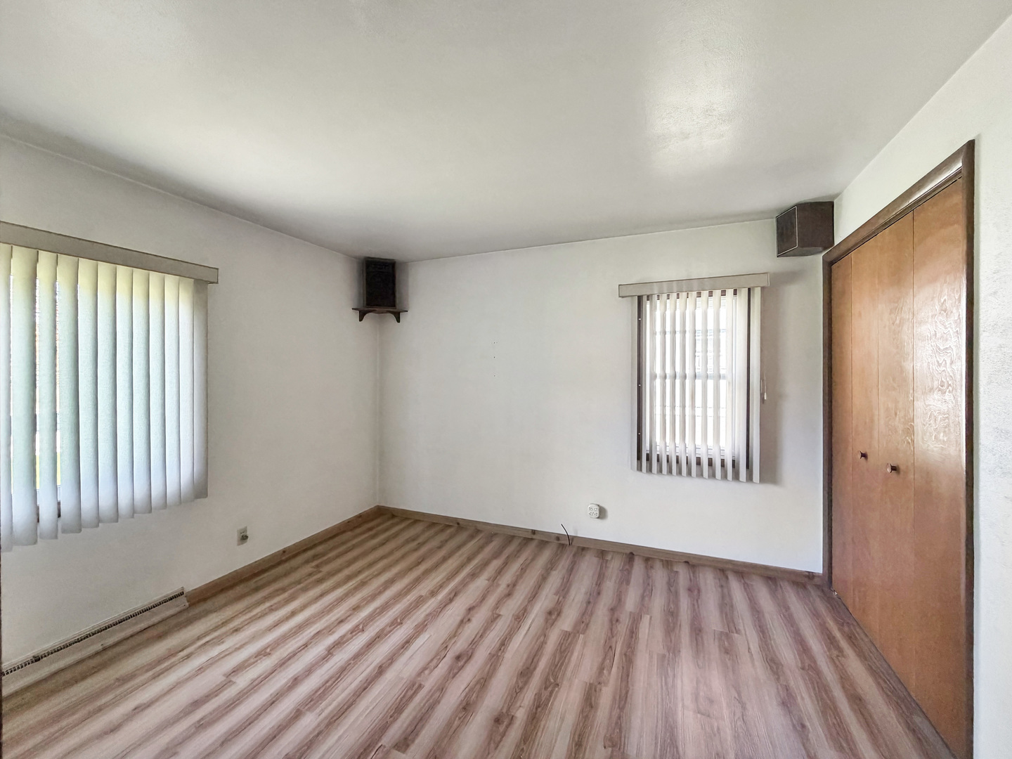 Empty bedroom with wood-look flooring, vertical blinds, and a large wooden closet.