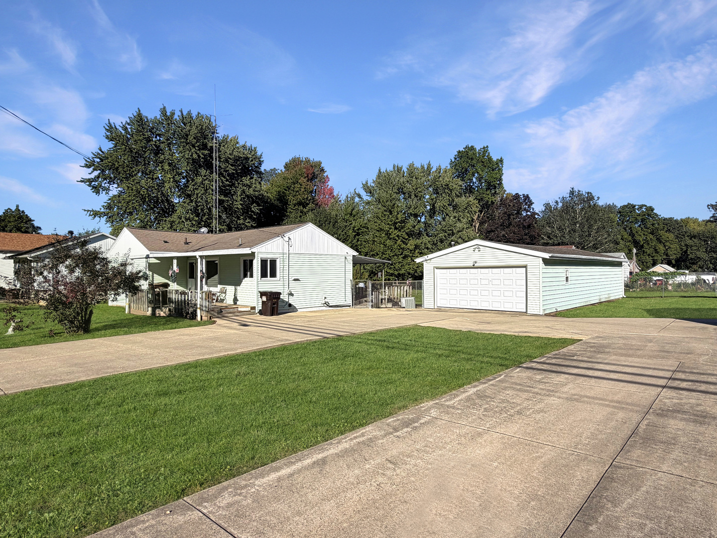 Single-story light-sided house with a small porch, detached garage, and broad concrete driveway.