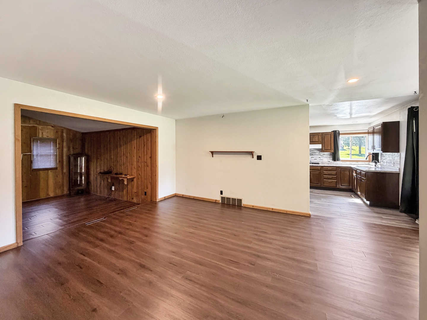 Open living area with dark wood flooring, a wood-paneled adjoining room, and a dark-cabinet kitchen.