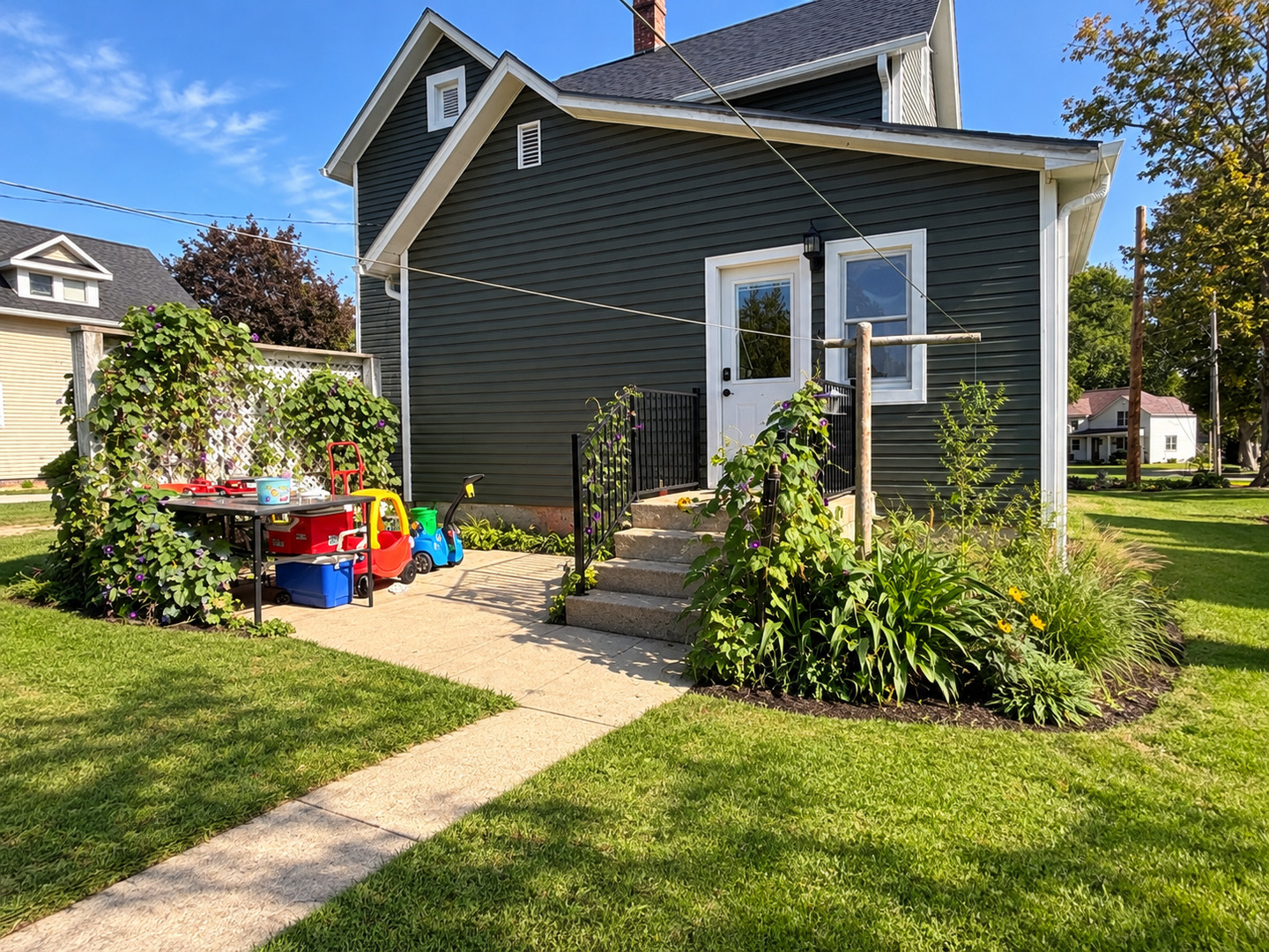 Green-sided house with a side entrance, garden beds, and a paved play area.
