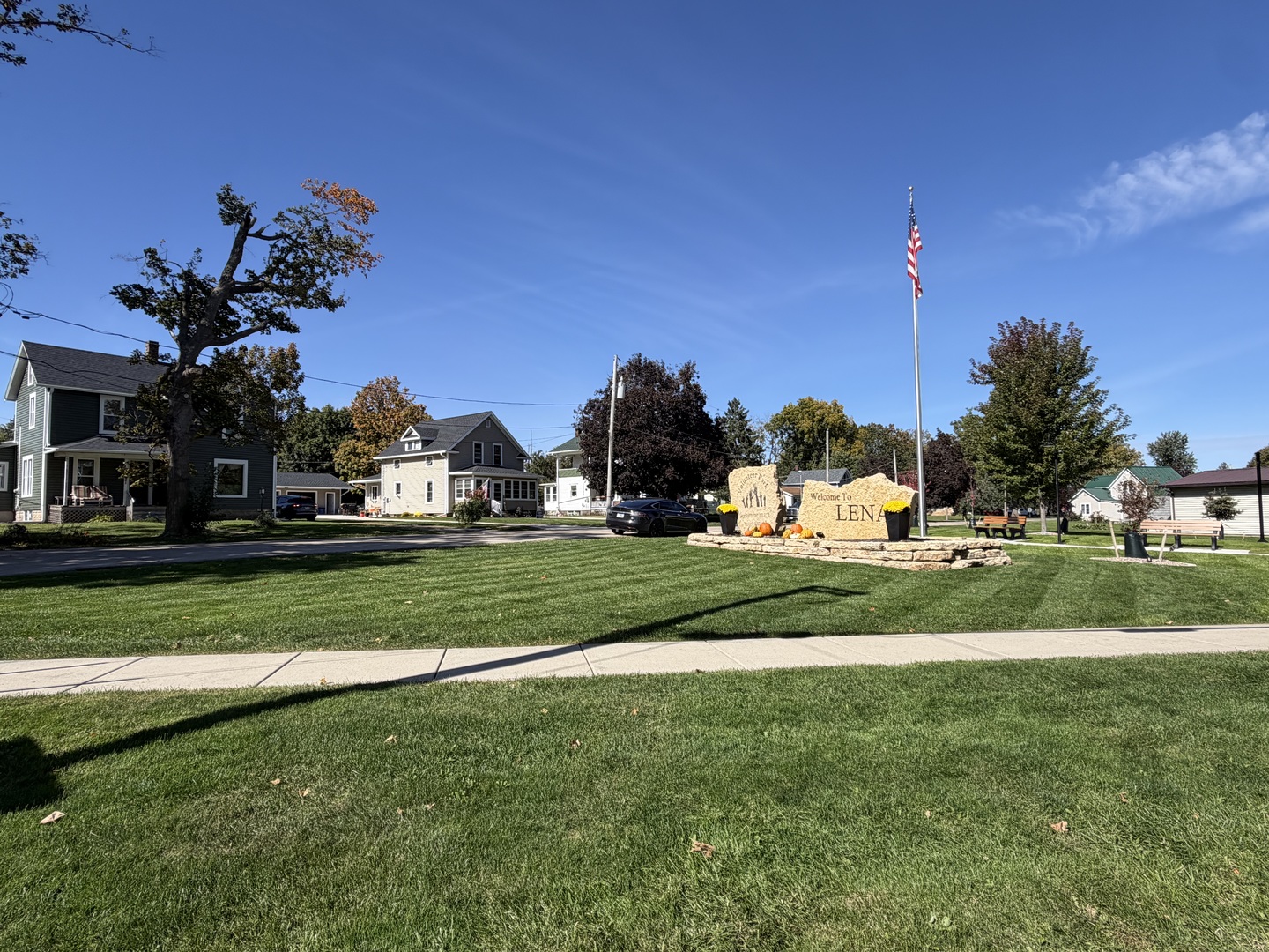 Welcome-to-Lena stone sign beside a flagpole and neighborhood street.