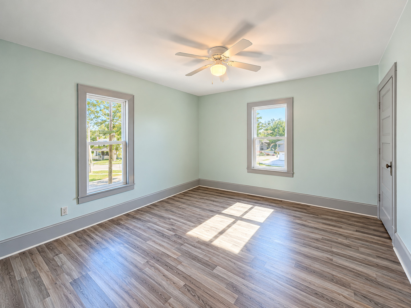 Empty room with pale green walls, two windows, wood-look flooring, and a ceiling fan.
