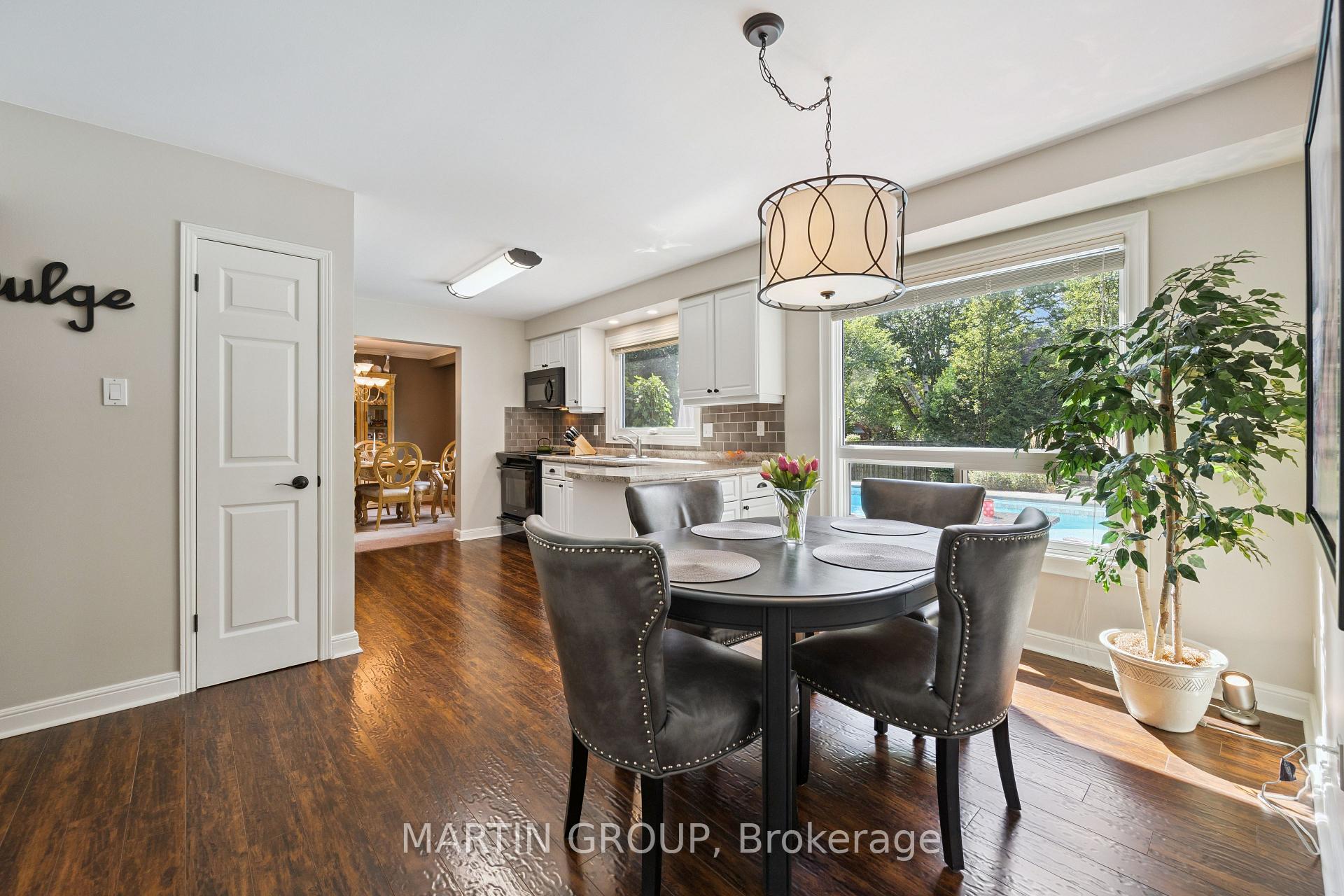 Dining area with a dark round table beside the kitchen and large windows overlooking a backyard pool.