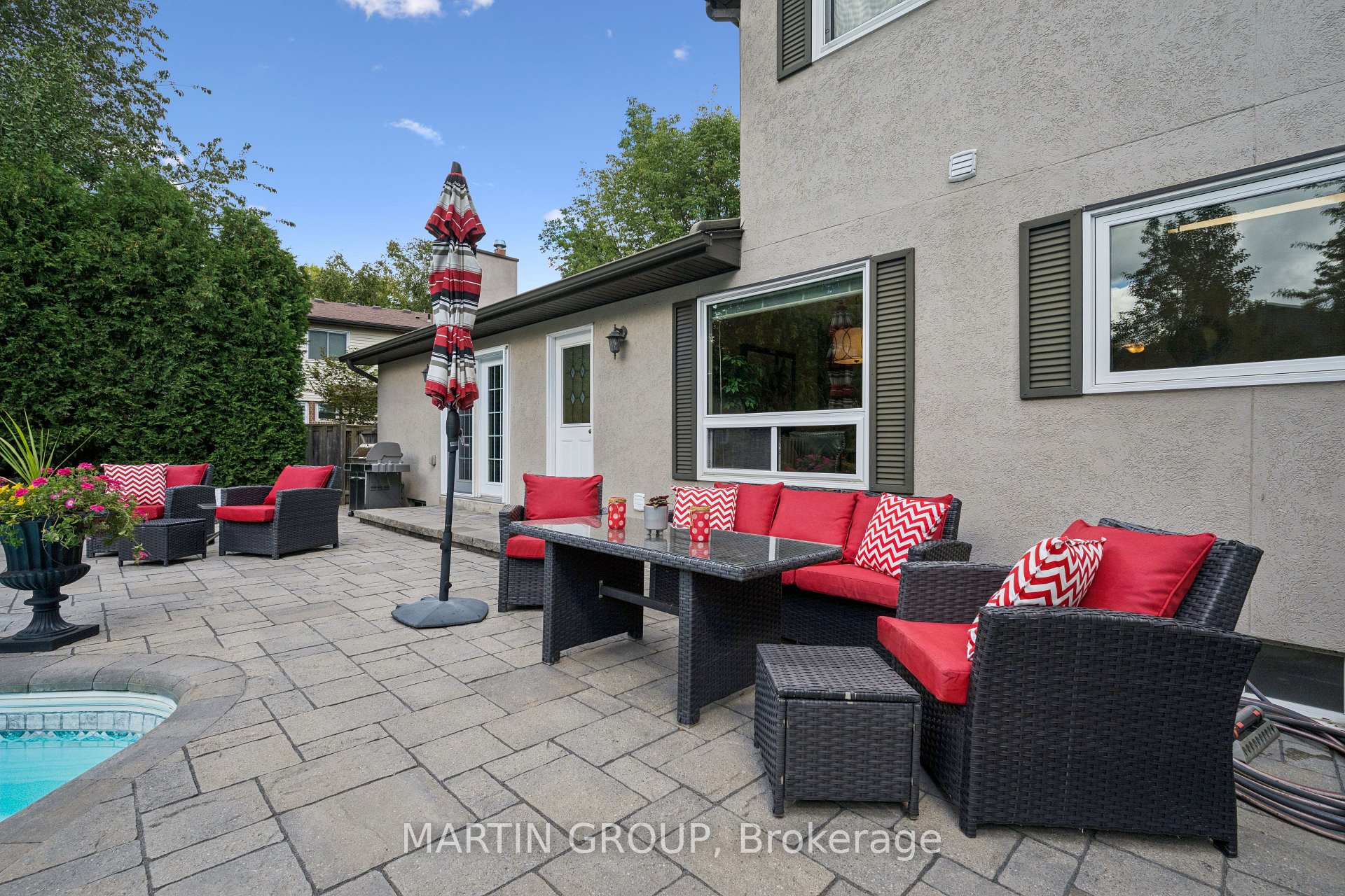 Backyard patio with red-cushioned wicker seating, a striped umbrella, and a swimming pool edge.