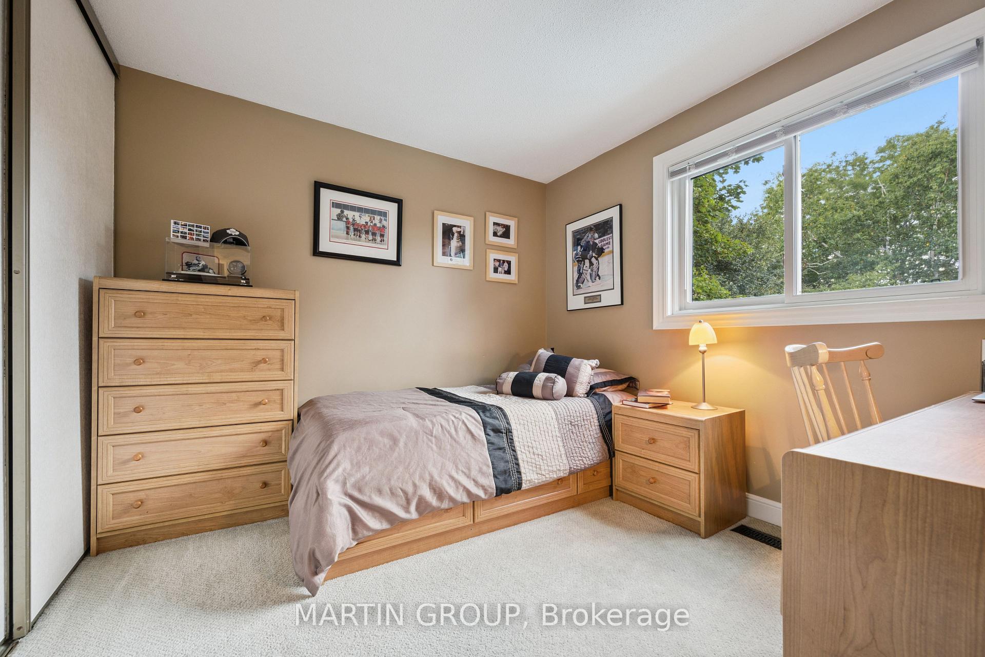 Furnished bedroom with wooden furniture, hockey memorabilia, and a large window overlooking trees.