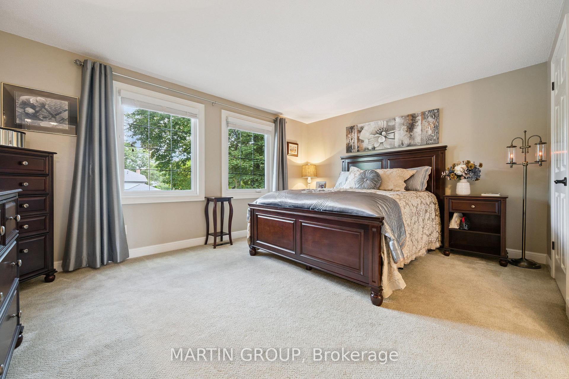 Furnished bedroom with a dark wood bed, neutral bedding, beige carpet, and two large windows.