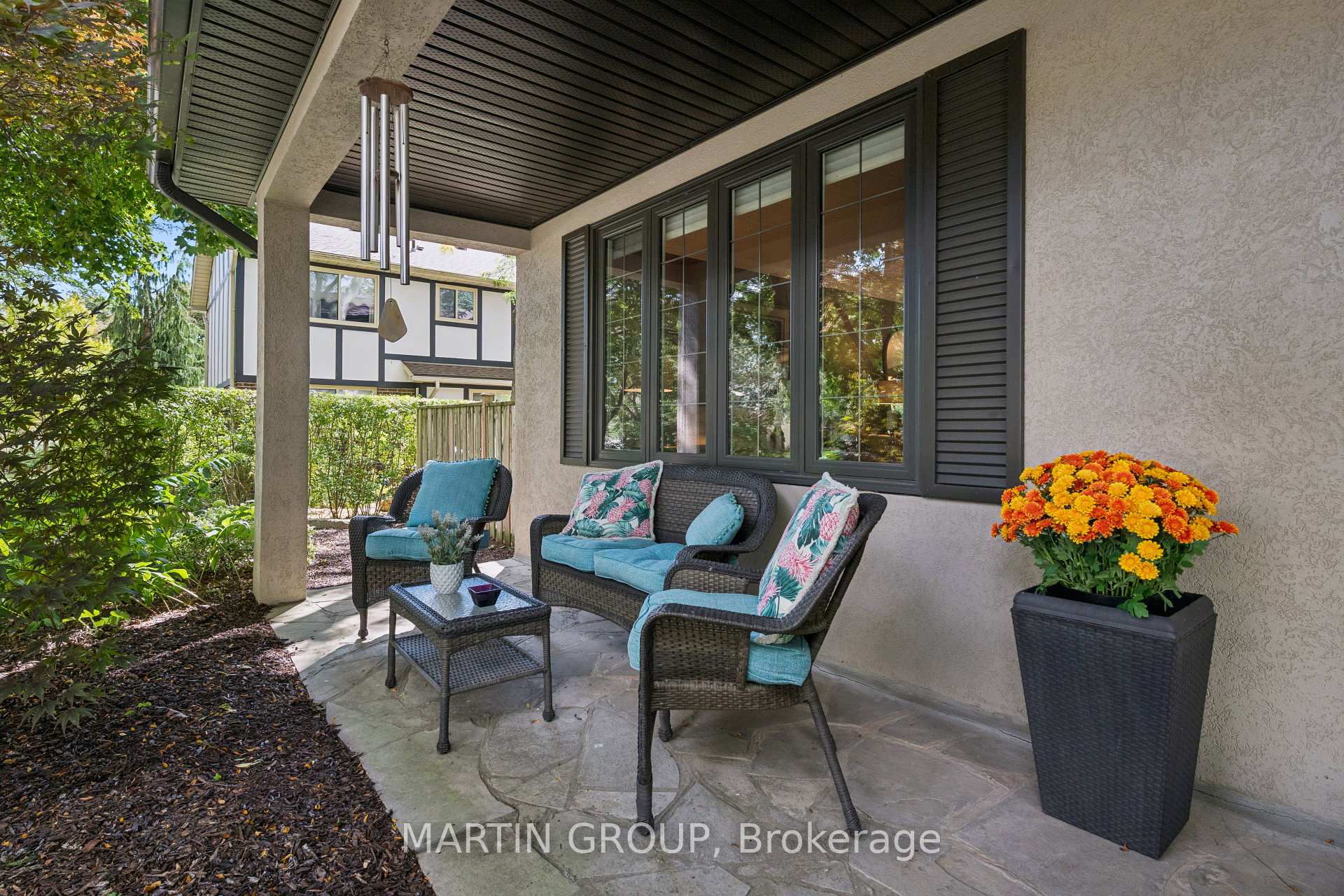 Covered stone patio with wicker seating, turquoise cushions, a hanging wind chime, and a flower planter.