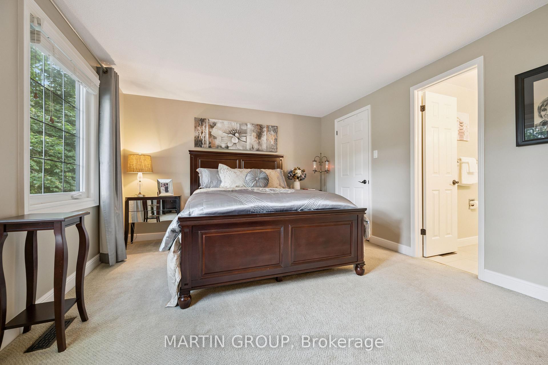 Furnished bedroom with a dark wood bed, silver bedding, and a large window overlooking leafy trees.