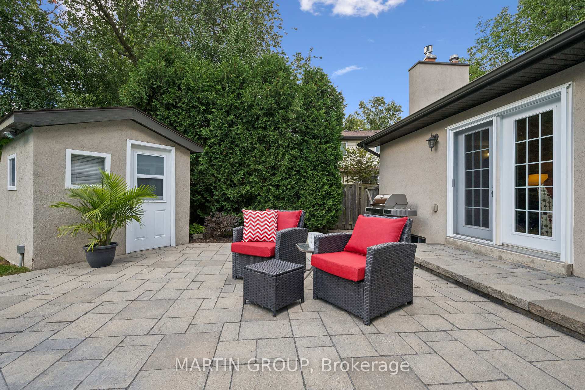 Backyard patio with red-cushioned wicker chairs, stone paving, and a barbecue beside the house.