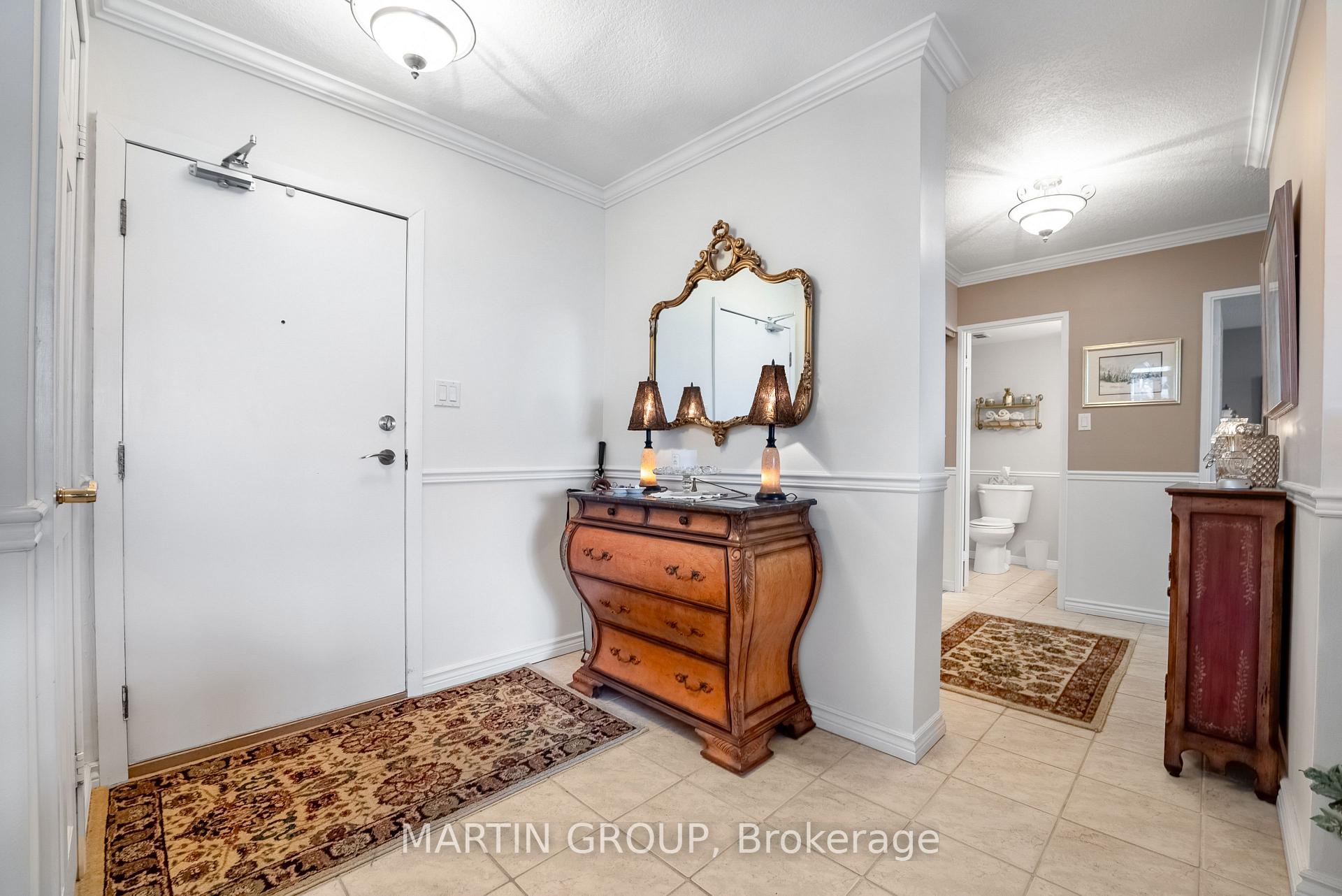 Apartment entry foyer with an ornate wooden chest, decorative mirror, patterned rugs, and an open bathroom doorway.