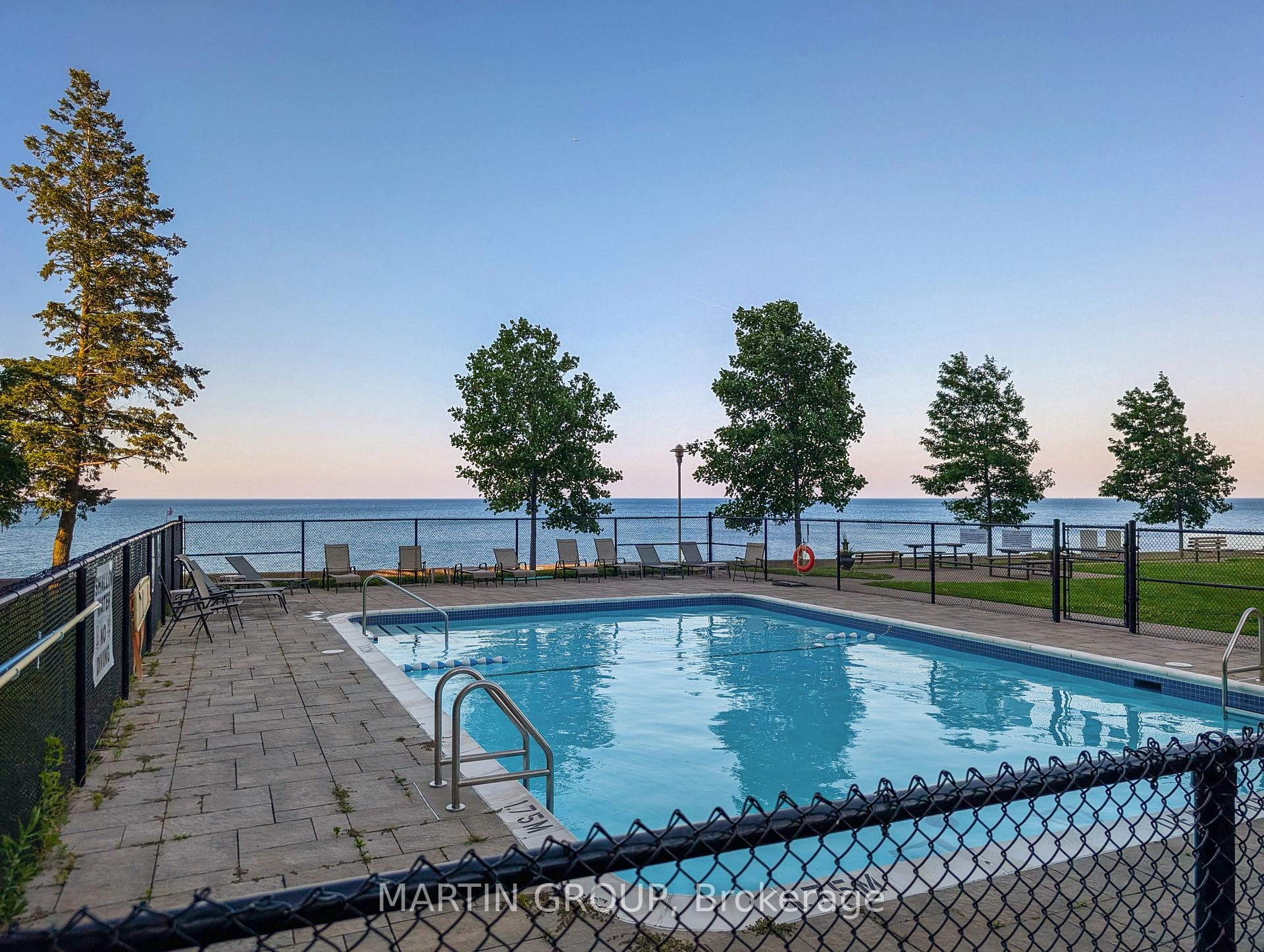 Outdoor pool beside a waterfront, with lounge chairs and trees along the shore.