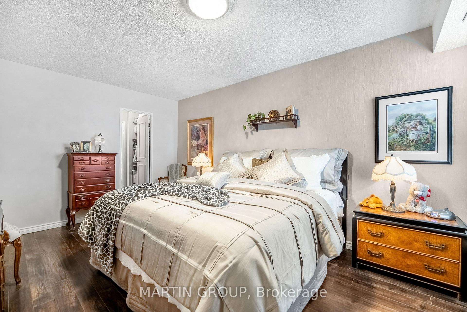 Bedroom with a large cream-bedded bed, wood dressers, framed artwork, and dark flooring.