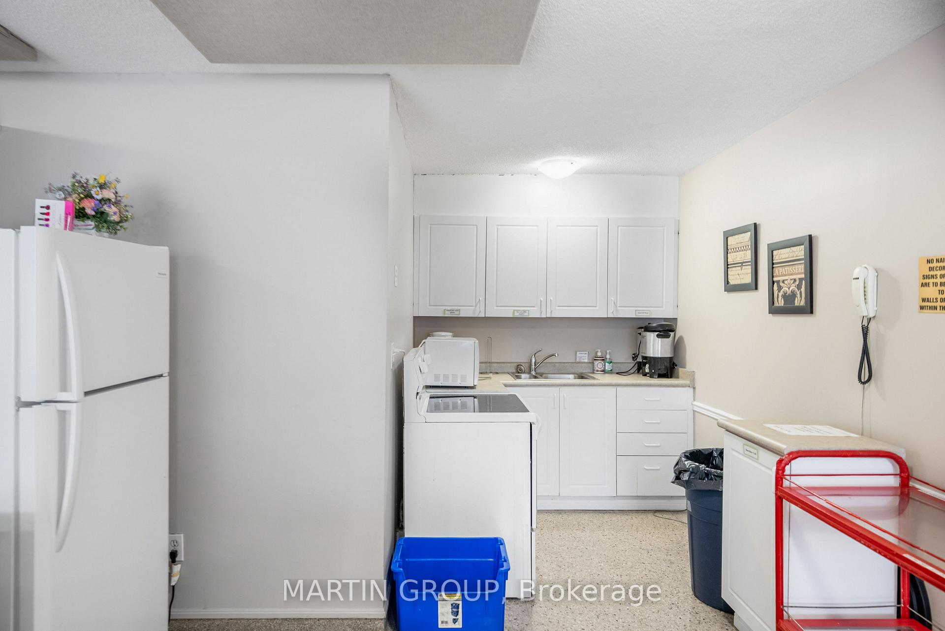 Kitchenette with white cabinets, refrigerator, stove, and sink; a blue recycling bin and red cart are in front.