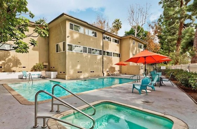 Residential courtyard pool and spa beside a tan multi-story building, with red umbrellas and turquoise chairs.