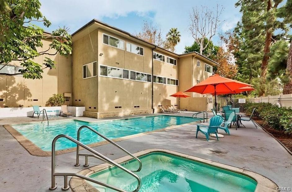 Residential building beside a swimming pool and spa, with red umbrellas and turquoise patio chairs.
