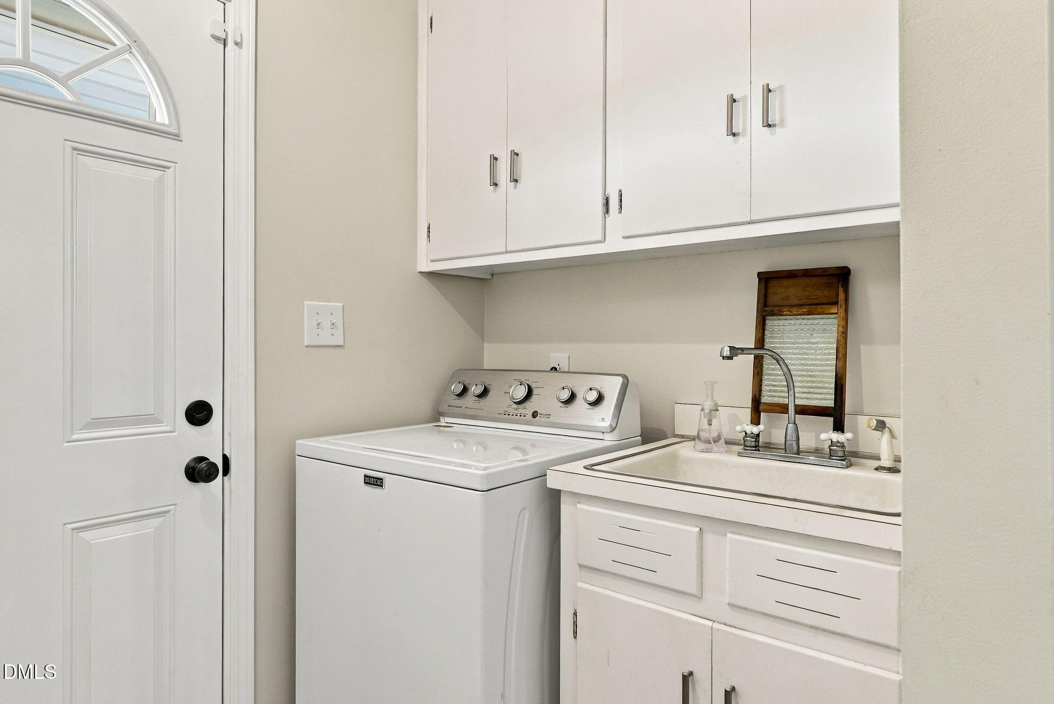 Laundry room with a top-loading washer, utility sink, and overhead cabinets beside a half-moon-windowed door.