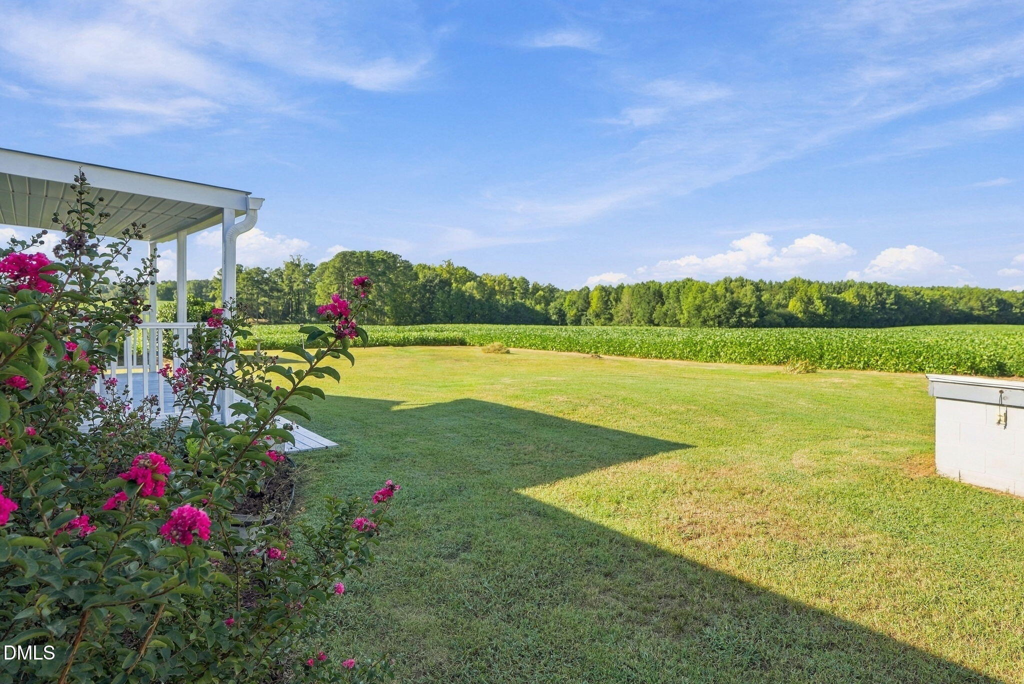 Covered porch beside a mowed lawn, pink flowering shrubs, and a green field backed by trees.