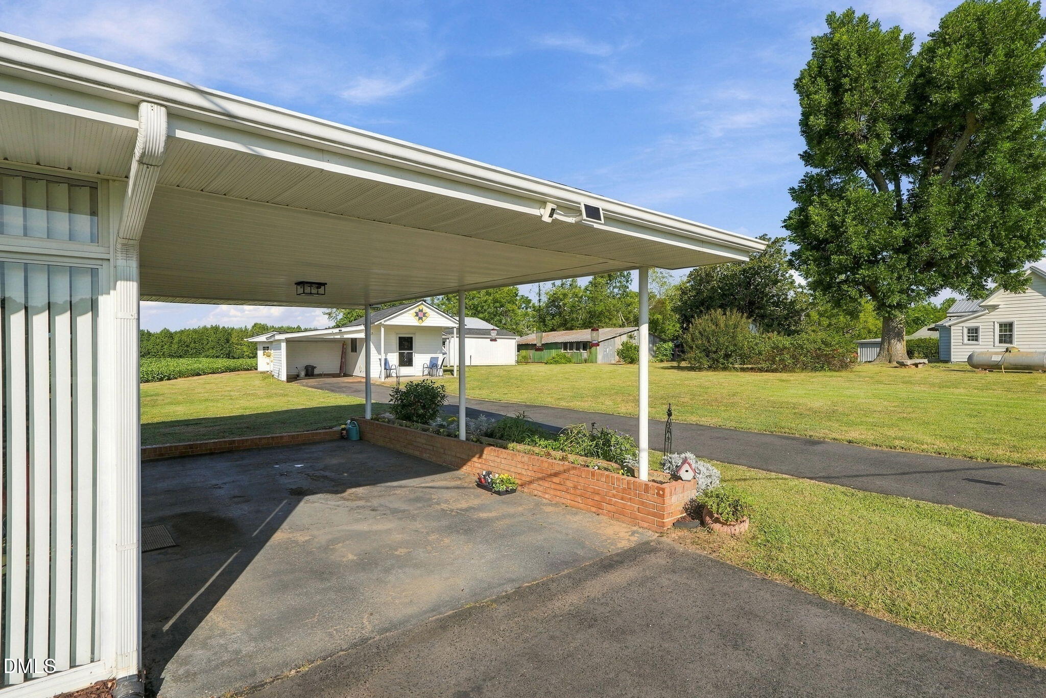 Covered carport and paved driveway overlooking a grassy yard with outbuildings and a large shade tree.