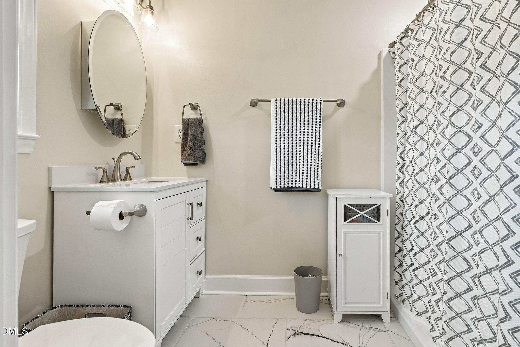Bathroom with a white vanity, oval mirror, patterned shower curtain, and marble-look tile floor.