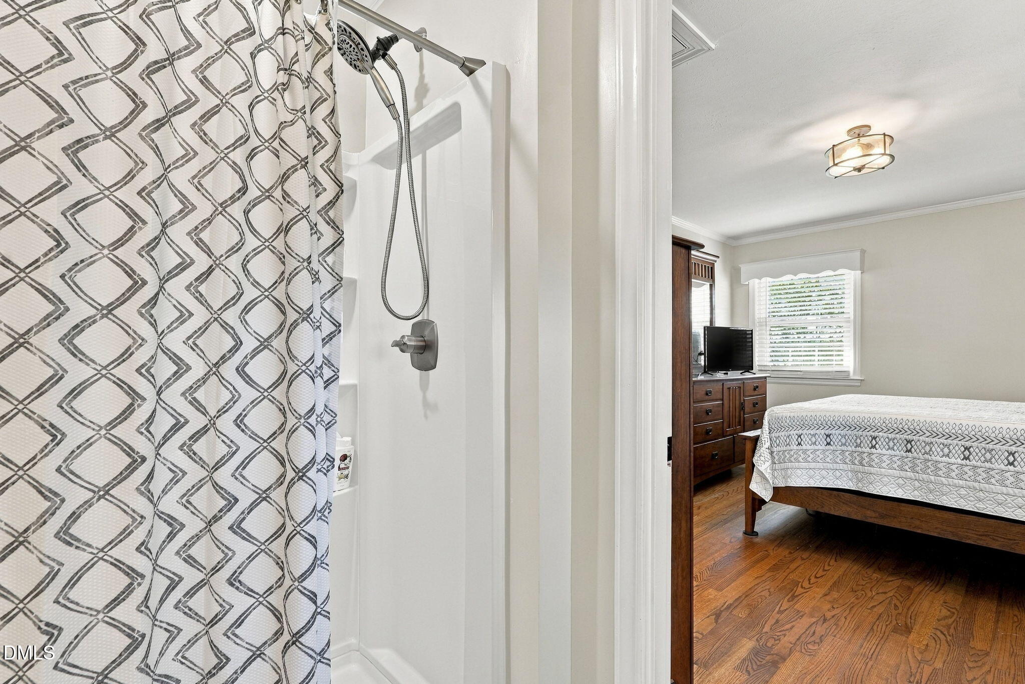 Bathroom shower with a geometric curtain overlooks a bedroom with hardwood floors and a wooden bed.