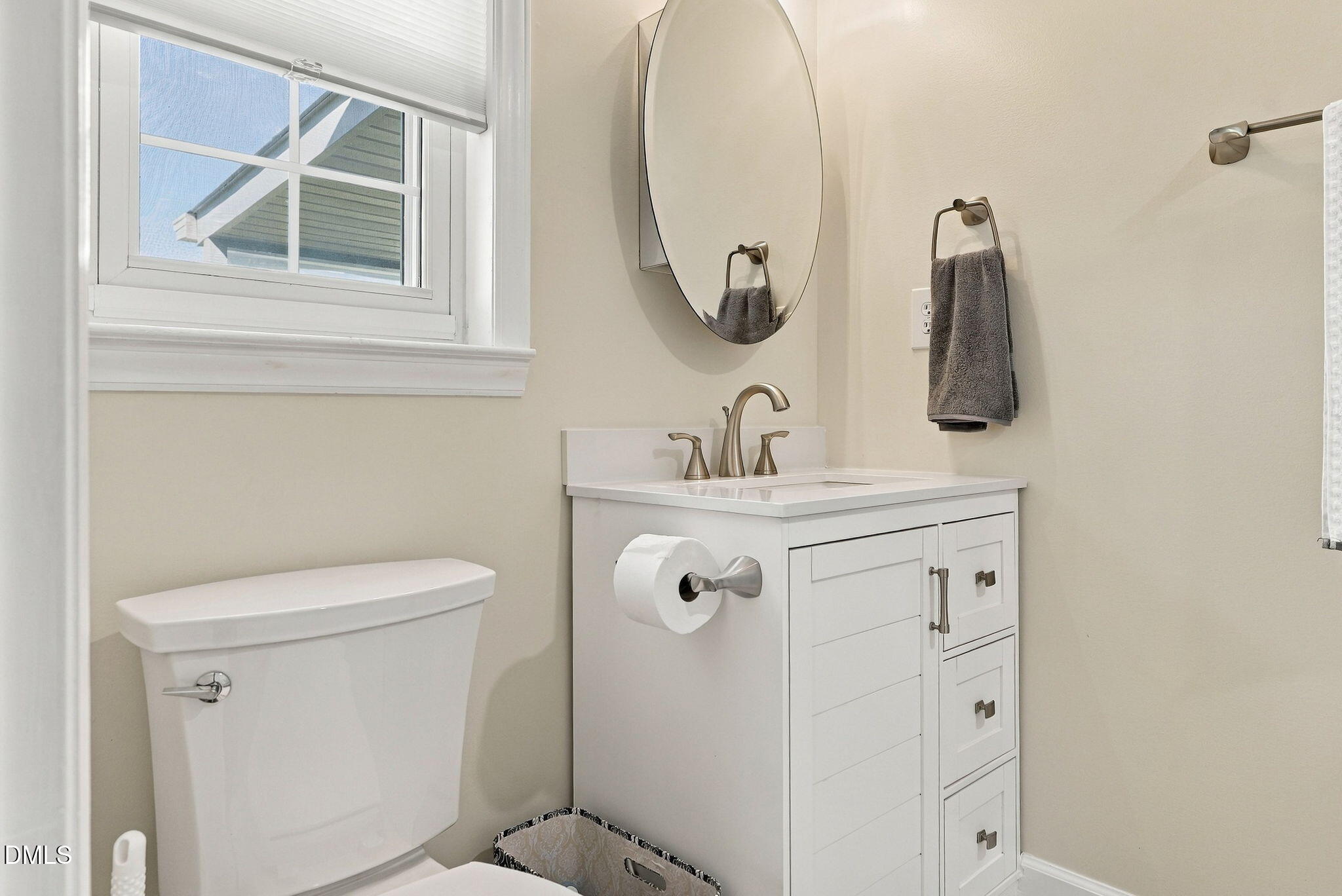 Bathroom with a white vanity, oval mirror, and window beside the toilet.