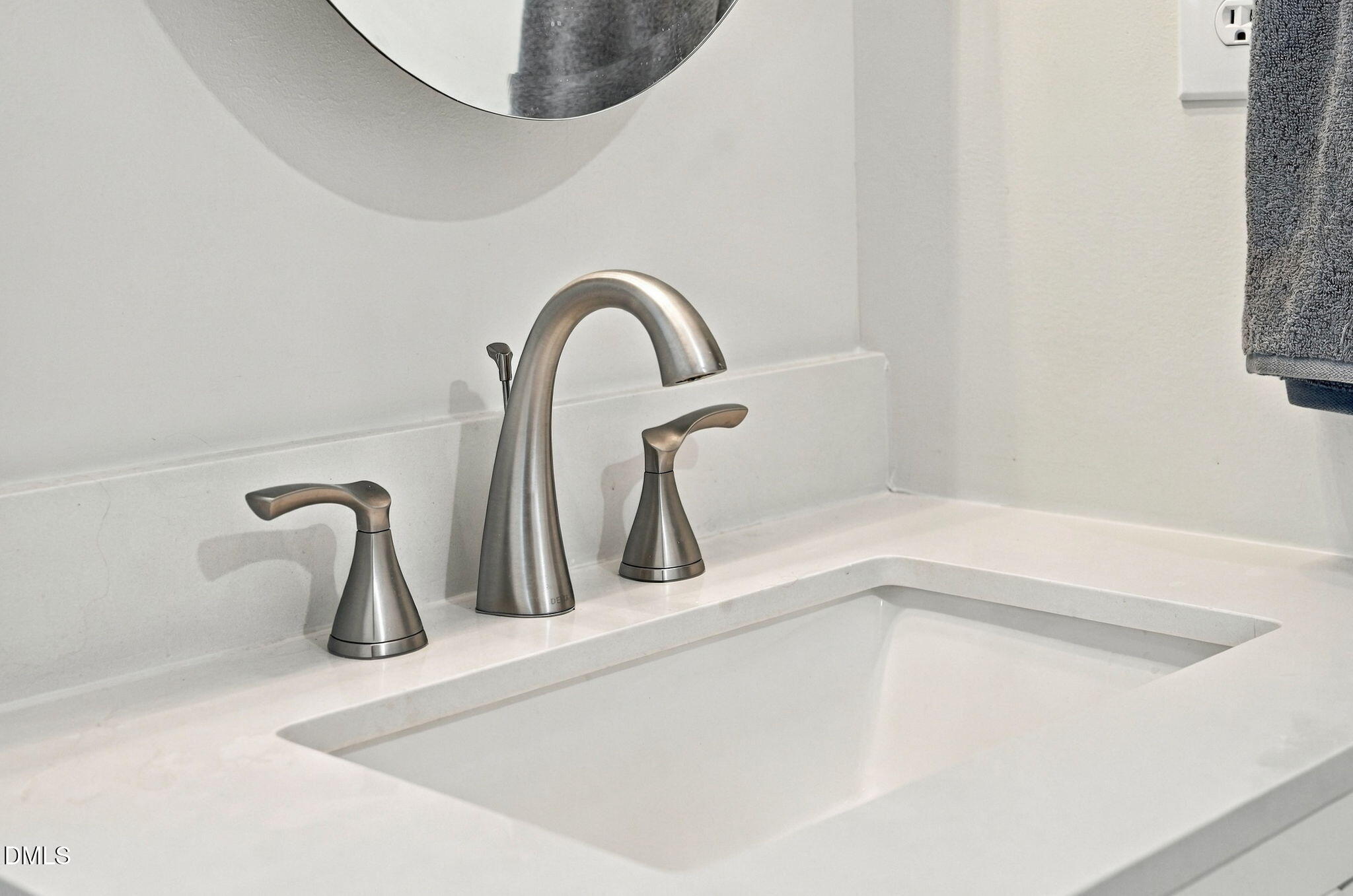 Bathroom sink with a brushed-nickel faucet, white countertop, partial round mirror, and gray towel.