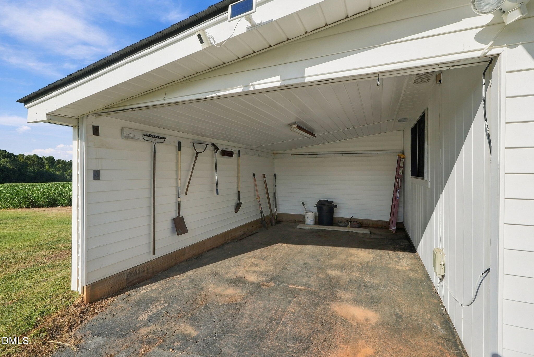 Attached white carport with gardening tools hanging on the walls and a cracked concrete floor.