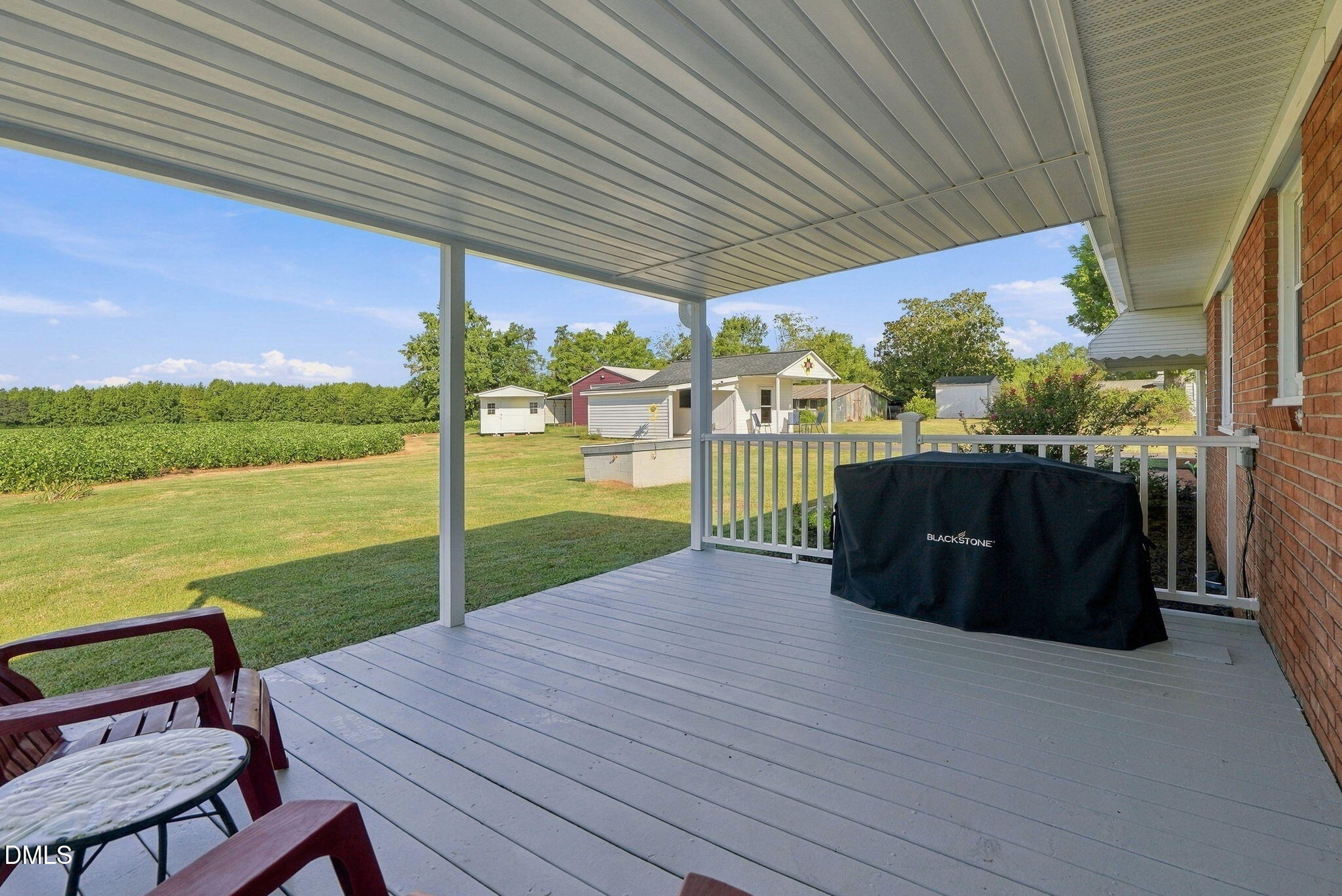 Covered deck with chairs and a grill overlooking a lawn, crop rows, and outbuildings.