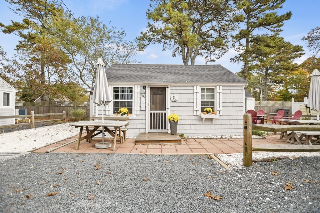 White-shingled cottage with flower boxes, screened entry, and a patio furnished with picnic tables and red chairs.
