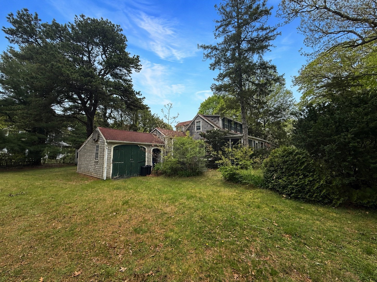 Shingled house and detached green-door garage amid mature trees, viewed across a broad lawn.