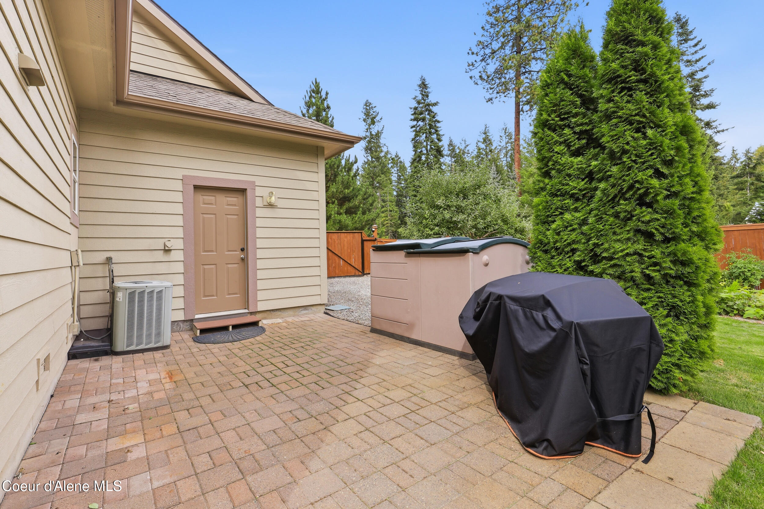 Paved patio with a hot tub, covered grill, and tall evergreens beside the house.