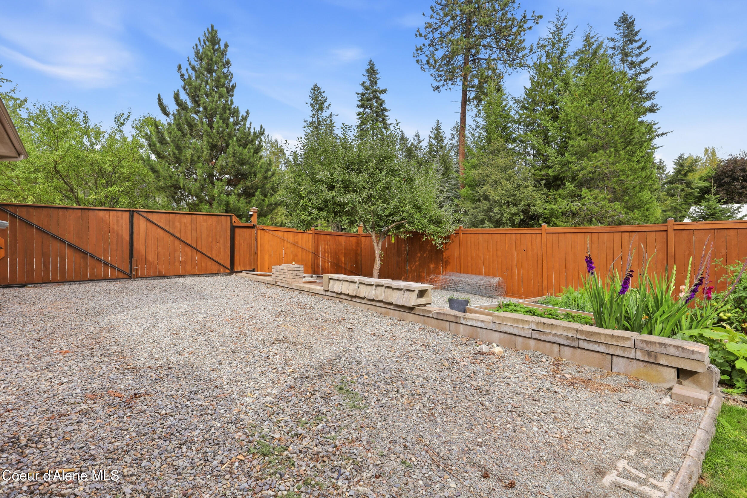 Fenced backyard with a gravel yard, raised garden beds, and purple flowers.