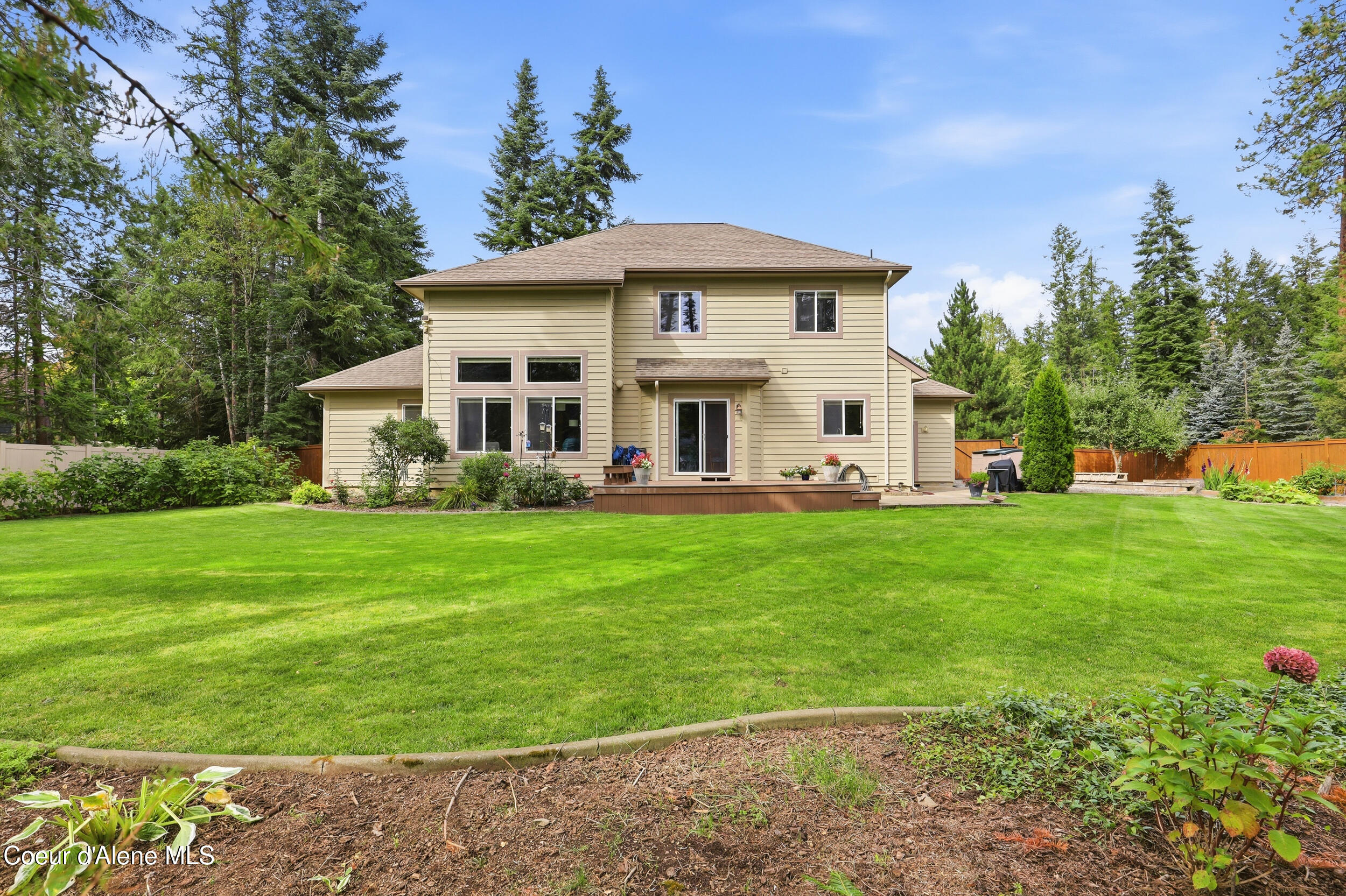 Two-story beige home behind a broad lawn, with a small deck and mature evergreens.