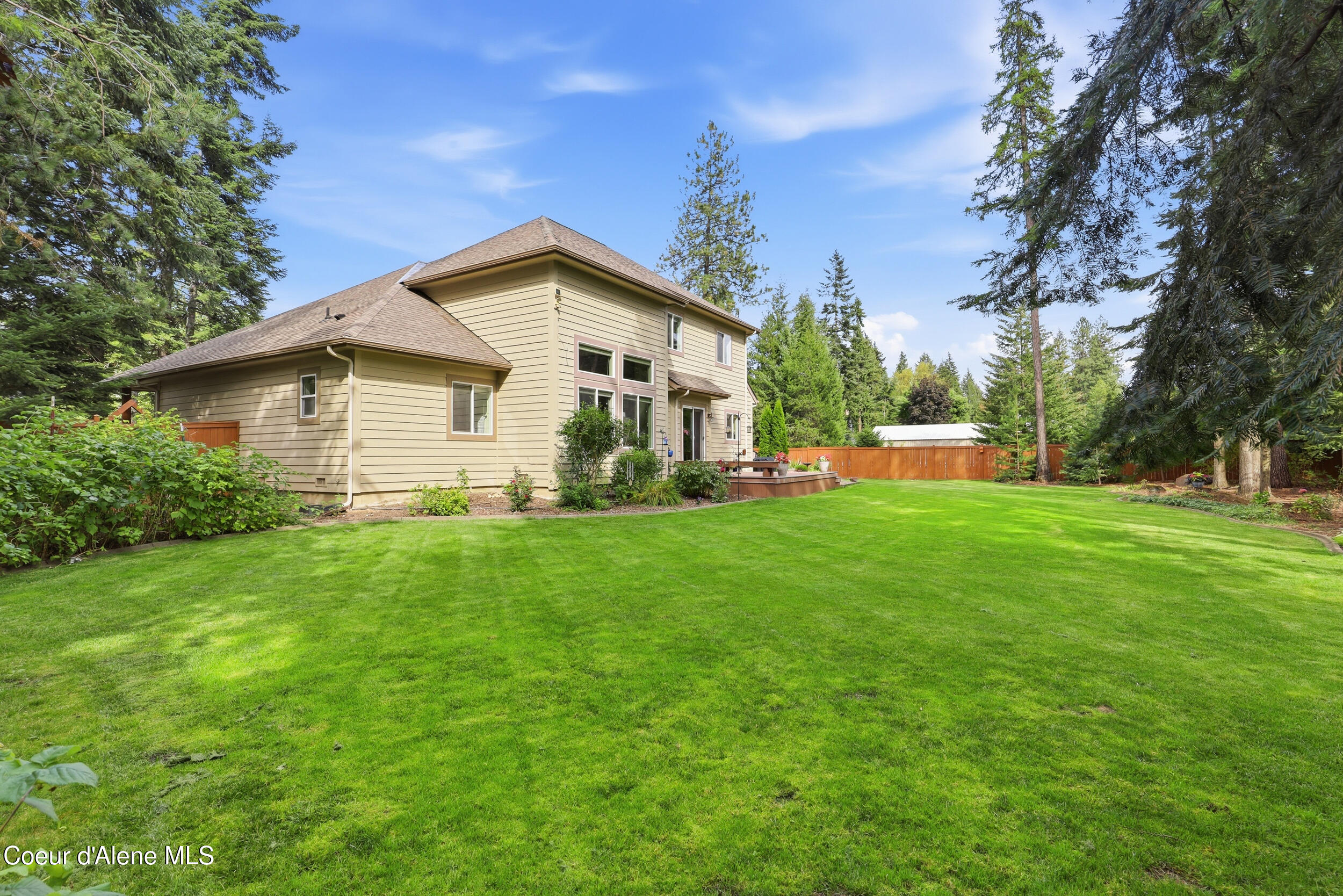 Rear of a two-story beige home with a raised deck, broad lawn, wooden fence, and mature evergreens.