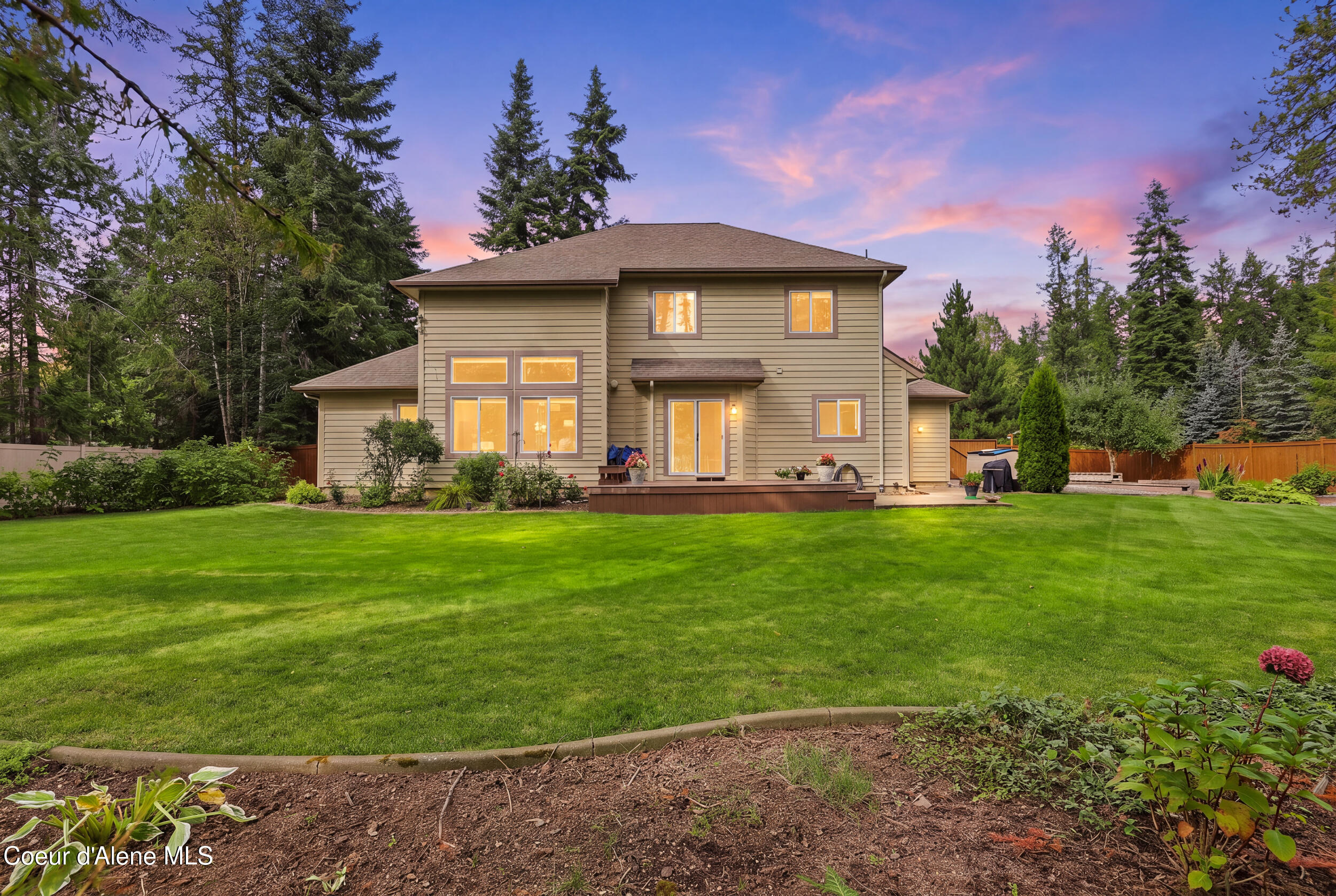 Two-story tan house with a small deck and broad lawn, framed by mature trees at dusk.