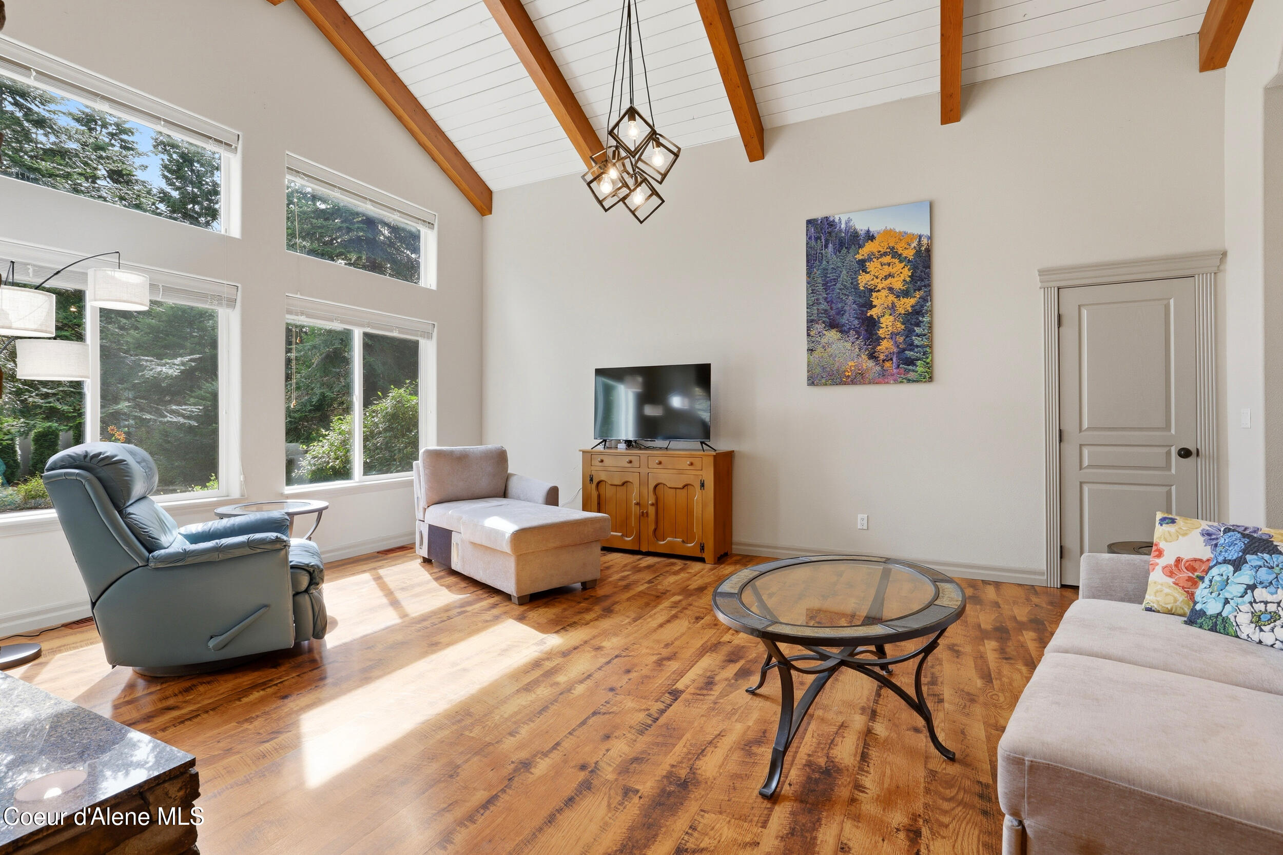 Vaulted living room with exposed wood beams, hardwood floors, and large windows overlooking trees.
