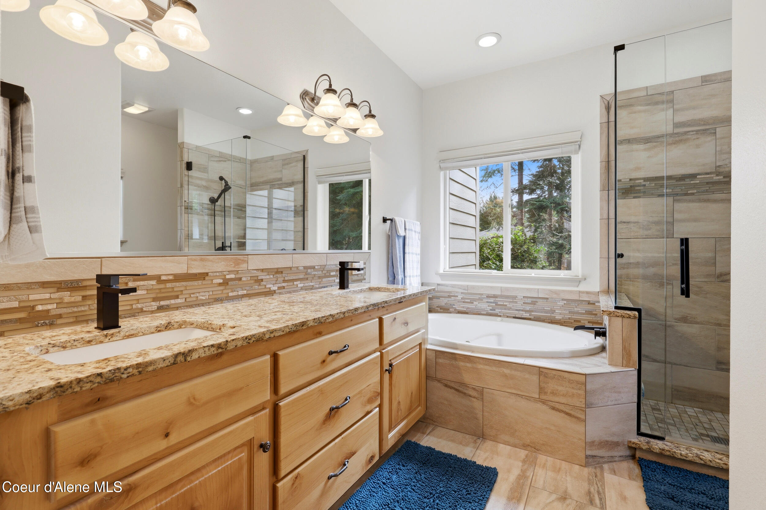 Bathroom with a long wood vanity, granite counter, tiled soaking tub, and glass shower beside a window.