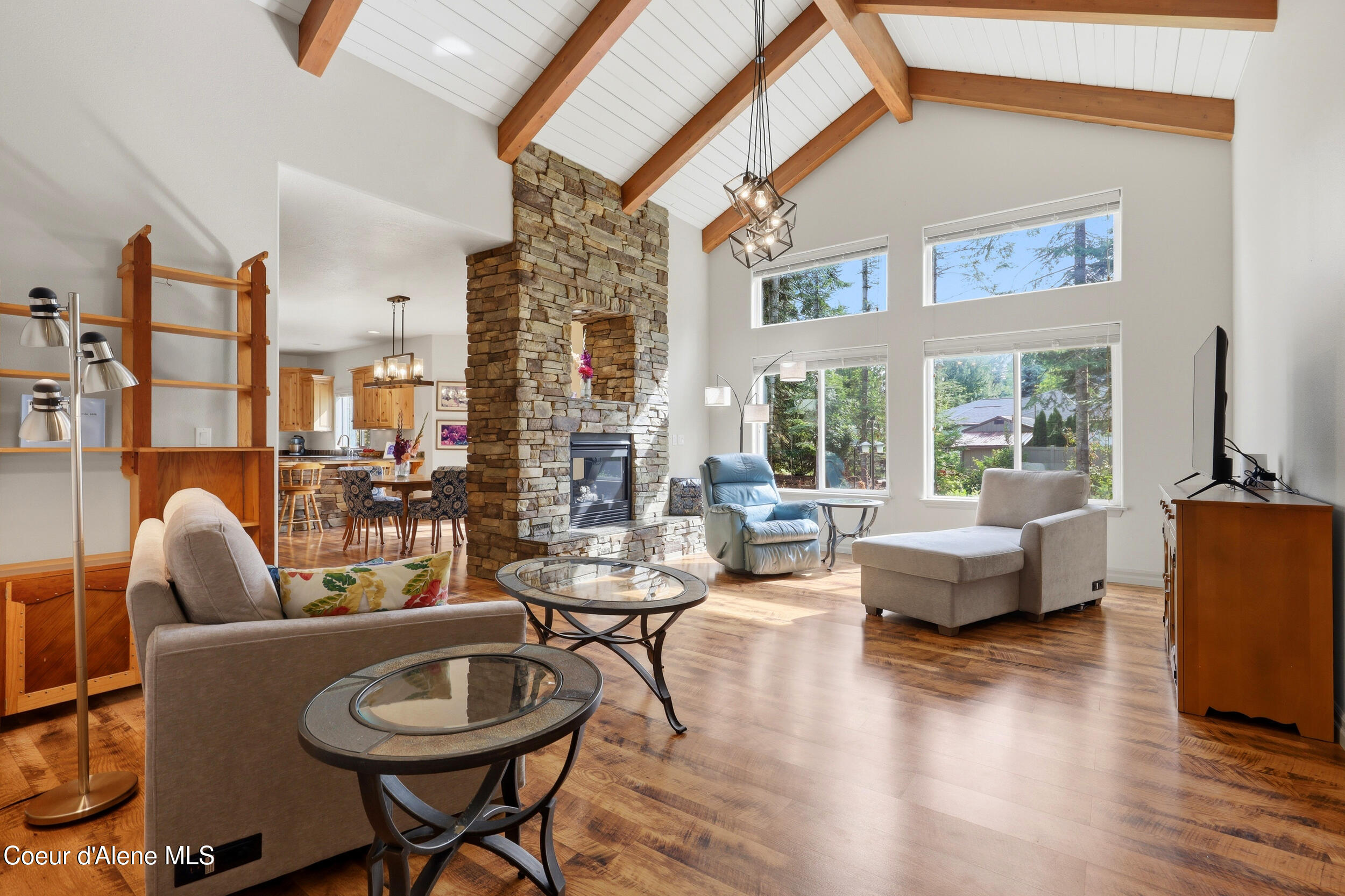 Vaulted living room with exposed wood beams, a tall stone fireplace, hardwood floors, and large windows.