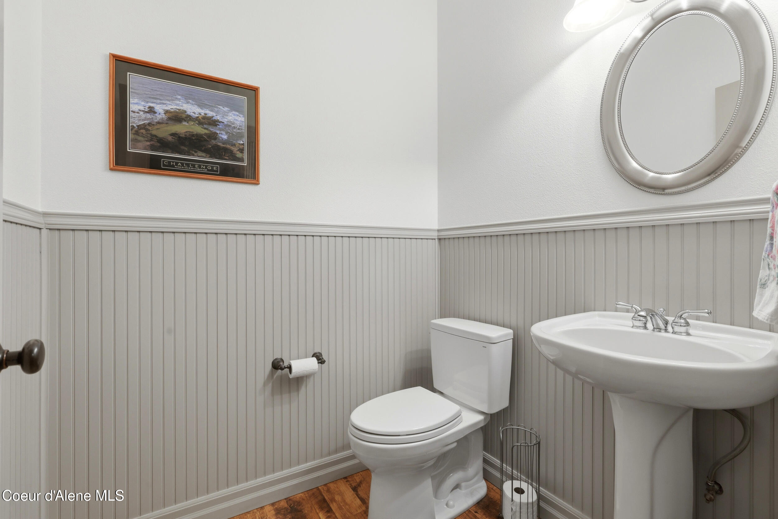 Half bathroom with a white toilet, pedestal sink, gray beadboard walls, and a large round mirror.